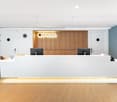 Large open office space with a modern and minimalist design. The reception area features a white curved desk with a wooden accent wall displaying the 'SPACES' logo. The space has hardwood flooring, white walls, and recessed lighting fixtures. There are tw