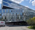 Large modern glass and steel commercial building with the name 'SPACES' prominently displayed. The building has multiple floors and a parking area in front. There are trees and landscaping around the building.