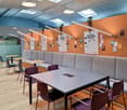 Wooden floor, tables and chairs, grey sofa, ceiling lights, orange wall with plant and hanging posters, windows, door