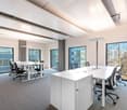 Spacious open-plan office with large windows providing a view of the surrounding trees and buildings. The room features white desks, black office chairs, and a conference table. The ceiling has exposed beams and lighting fixtures. The floor is covered in