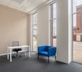 The image shows a modern office space with a white desk, a blue armchair, and large windows overlooking the exterior. The room has a neutral color scheme with beige walls and gray carpeting. The overall design appears to be minimalist and professional.