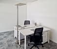 White desk with a computer, office chair, and a floor lamp. The room has white walls and a gray carpet floor.