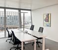 This image shows a modern office space with large windows providing a view of the surrounding buildings. The room has a minimalist design with white walls, a gray carpet floor, and a long rectangular table with black office chairs. There is also a framed