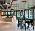 Open office space with large windows, high ceilings, and exposed ductwork. Includes a reception desk, meeting table with stools, and potted plants throughout the space.
