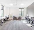 This image shows a modern and well-lit office space. The room has large windows that provide natural light, and the walls are painted in a neutral gray color. The space is furnished with several desks and office chairs, as well as a couple of comfortable