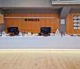 The image shows a modern office space with a large wooden reception desk featuring the 'Spaces' logo. The space has a minimalist and contemporary design, with gray walls, wooden accents, and a display of various office supplies and decor items on the desk