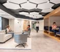 people sitting on couches and chairs, table with laptop, man walking, hexagonal ceiling panels, warm-toned wooden partitions, wooden floors