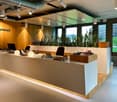 Modern office reception with wooden desk, office chairs, monitor, books, and a large window overlooking a garden