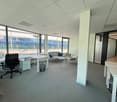 Spacious open office layout with glass walls, modern furniture, office desks, chairs, glass coffee table, couch, trash can, carpeted floor, bright lighting.