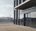 Modern building with glass facade, entrance with automatic sliding doors, name board reading 'Grosspetertower', metal railings, street lights, and a fence in the background.