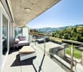 Spacious balcony with glass railing, offering a scenic view of the surrounding hills and greenery. The balcony features comfortable outdoor furniture, including a sofa and a wooden bench, providing a relaxing space to enjoy the natural surroundings.