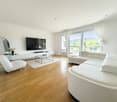 Spacious living room with hardwood floors, large windows providing a scenic view, white leather sofas, a TV mounted on the wall, and a gold-framed mirror on the side. The room has a modern and minimalist design aesthetic.