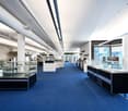 Large open space with blue carpeted floor, various kitchen and home appliances on display, including dishwashers, ovens, and refrigerators. The space has high ceilings with exposed ductwork and large windows providing natural light.