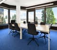 Large conference room with floor-to-ceiling windows, white table, black office chairs, blue carpeted floor, and views of trees and buildings outside
