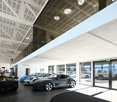 Garage showroom with multiple cars, elevated walkway, glass walls and ceiling, illuminated lights.