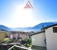 The image shows a view of a town or village nestled in the mountains, with a lake or body of water in the background. The buildings appear to be residential, with a mix of colors and architectural styles. The Assofide S.A. logo is visible at the top of th