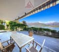 Outdoor patio with wooden table and chairs, overlooking a scenic lake and mountains in the background. The patio has a covered awning and is surrounded by lush greenery.