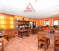 The image shows the interior of a restaurant or cafe. The space has an orange color scheme with wooden furniture, including tables and chairs. There is a counter area with shelves and decor items. The ceiling has recessed lighting fixtures. The overall at