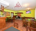 The image shows the interior of a restaurant or bar. The space has yellow walls, wooden furniture including tables and chairs, and a fireplace. There is a bar area with shelves displaying various alcoholic beverages. The room also has a TV mounted on the