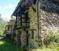 A rustic stone building with a wooden balcony or terrace, surrounded by lush greenery and mountains in the background.