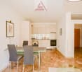 The apartment has an open-plan dining and living area with a modern kitchen, wooden floor, a dining table with four chairs, a glass top, and a glass side table. There is a picture frame on the wall, a radiator, and an entrance to another room. The kitchen