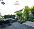 Outdoor seating area with a stone bench, a table, a small garden with flowers, plants, and a stone wall. There is a house in the background.