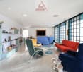 modern living room with white walls, gray floor, large windows, various chairs and a sofa, a TV, and decorative items on shelves