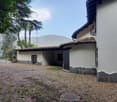 Two-story house, white walls, brown tiled roof, brick driveway, iron gate, black window shutters, fenced garden, mountainous landscape in the background
