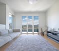 Spacious living room with large windows offering a scenic view of the city and mountains, hardwood floors, gray sofa, TV stand, and decorative plants.