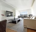Spacious living room with large windows offering a scenic view, hardwood floors, beige sofa, black coffee table, TV mounted on the wall, and decorative elements such as a vase of flowers.
