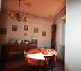 The image shows a cozy and traditional dining room with a chandelier, ornate ceiling, framed artwork on the walls, and a wooden cabinet or sideboard. The dining table is set with a patterned tablecloth and a decorative centerpiece. The room has an overall