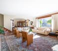 Spacious living room with large windows, hardwood floors, ornate area rug, wooden furniture, and a fireplace. The room has a cozy and elegant feel with potted plants and decorative items adding to the ambiance.