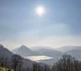 The image shows a scenic mountain landscape with a lake in the foreground. The sun is shining brightly in the sky, casting a warm glow over the scene. The mountains in the distance are partially obscured by haze, creating a sense of depth and atmosphere.