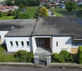 Two-story modern house with a flat roof, white exterior walls, and yellow accents. The house has a garage and is surrounded by a well-maintained garden with trees and shrubs. The property is situated on a hill overlooking a residential neighborhood.