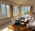 Large windows, curtains, white walls, wooden table, couch, carpet, radiator, framed picture, lamp