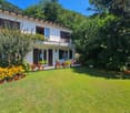 2 story house, white exterior, wooden doors, balcony with railing, flower-filled yard, well-manicured lawn, multiple flower pots