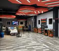 Interior of a car wrap shop, featuring red octagonal ceiling lights, large tables with computers, storage units, and tools, a monitor displaying an image, office chairs, and shelves with items.