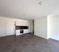 The image shows a modern, open-plan apartment with a kitchen and living area. The apartment has white walls, gray tile flooring, and built-in white cabinets and appliances in the kitchen. The space appears to be well-lit with recessed lighting fixtures in