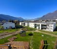 The image shows a modern apartment complex with several multi-story buildings surrounded by a well-maintained garden area. The buildings have balconies and the complex appears to be situated in a scenic location with mountains in the background.