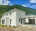 Two-story modern house with a white exterior, large windows, and a covered parking area. The house is situated in a scenic mountainous area with lush green vegetation surrounding it.