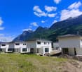 A row of newly constructed white houses with slanted roofs, surrounded by a grassy area and set against a backdrop of lush green mountains and a blue sky with fluffy white clouds.