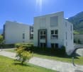 A modern, multi-story building with a minimalist, rectangular design. The building has large windows, a paved walkway, and a grassy area surrounding it. The building appears to be situated in a mountainous, forested area with a clear blue sky overhead.
