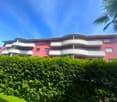 A multi-story apartment building with a red and white exterior, surrounded by lush greenery and palm trees. The building has curved balconies and large windows.
