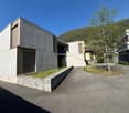 Modern concrete house, two-story, with a driveway, stone wall, and a tree in the front yard.