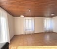 The image shows a spacious living room with a wooden ceiling, beige walls, and tile flooring. The room has large windows with sheer curtains, providing natural light. There is a light fixture hanging from the ceiling and the room appears to be empty, read