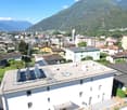 Aerial view of an apartment building with solar panels on the roof, surrounded by various other buildings, green vegetation, and mountains