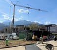 An ongoing construction site with multiple machinery vehicles, workers, a large crane, and nearby buildings, all under a clear blue sky.