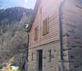 A rustic house with wooden sidings, a stone wall, a roof with a chimney and a wooden entrance.