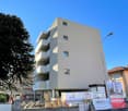 Newly constructed apartment building with multiple floors, concrete walls, and balconies. It has an elevated ground floor and is surrounded by a fence, trees, and construction vehicles.