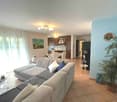 Large L-shaped sofa, white and blue cushions, glass table, round side table, potted plant, open view of kitchen with dining table and chairs